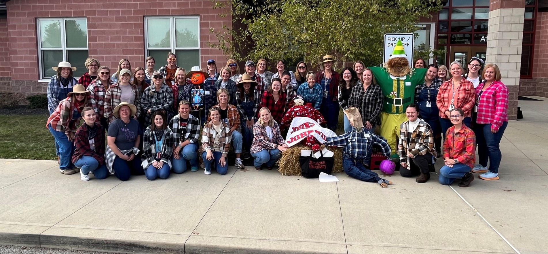 Elementary staff members dress in flannel and farm apparel for Dress for a Farmer Day, posing with scarecrows in front of elementary school