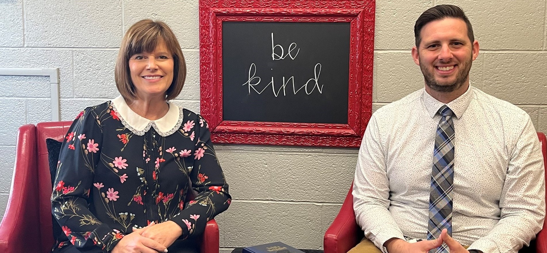 JMS Principal Mrs. Kris Almendinger & Dean of Students Nick Hancock seated in principal&#39;s office with framed message of &#34;Be Kind&#34; on the wall between them.