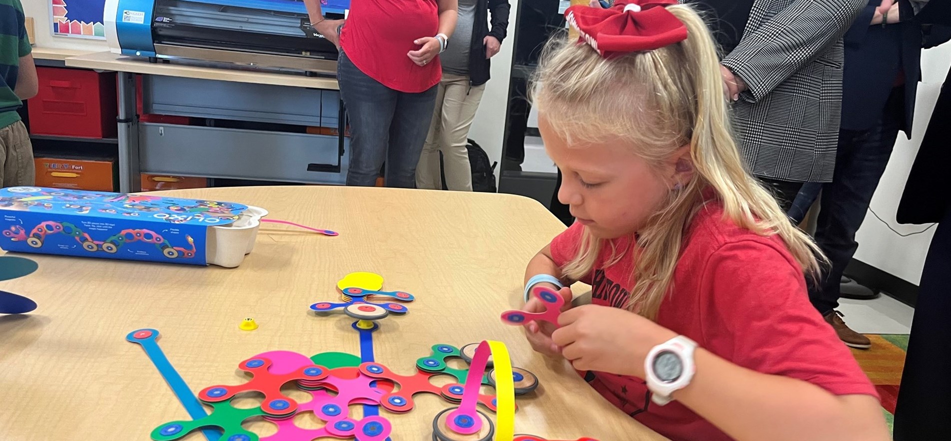 Elementary student in red shirt with red bow practicing STEAM exercise with craft parts at a table.